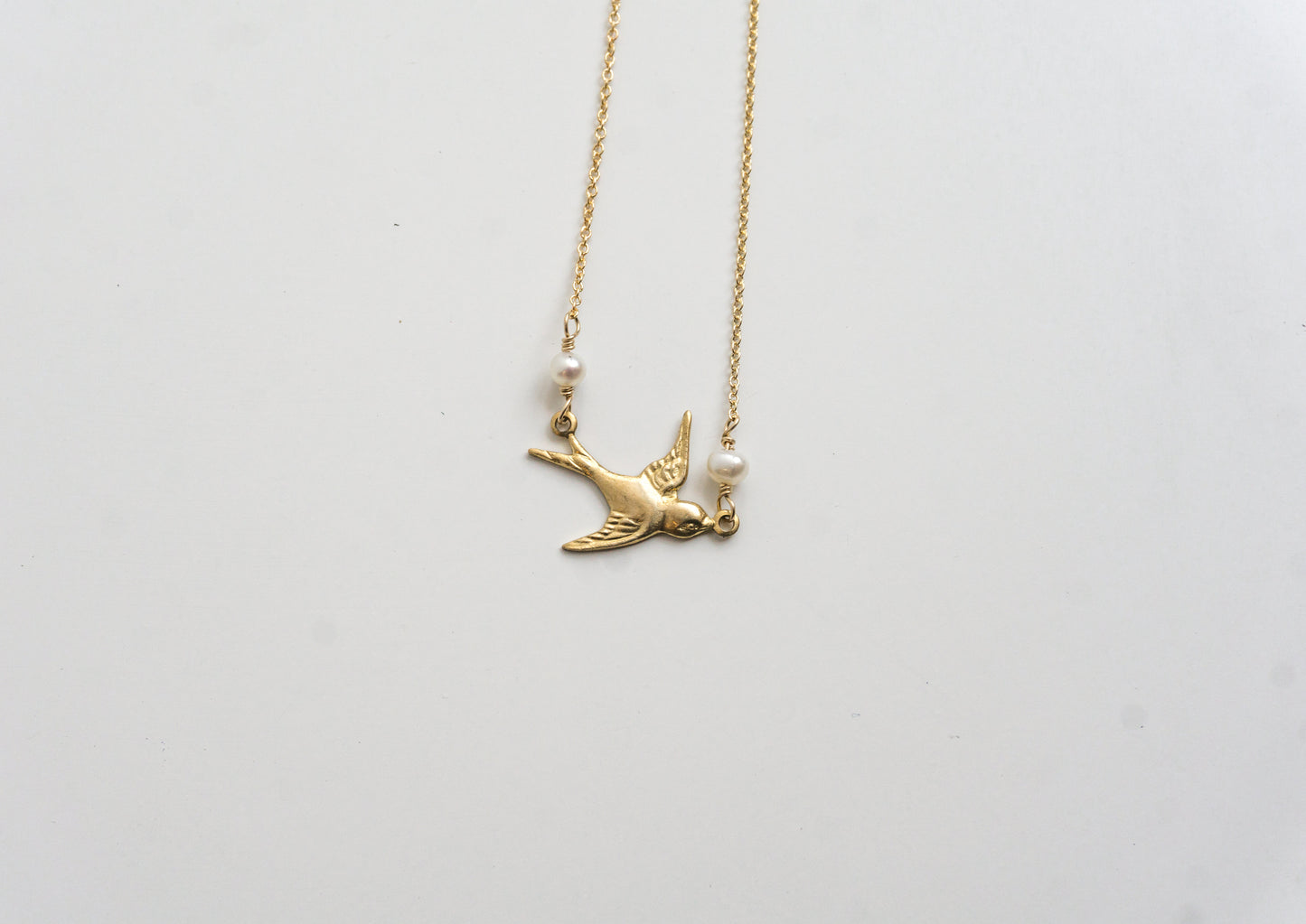 A gold-colored necklace with a charm resembling a bird and two freshwater pearls, displayed against a white background.