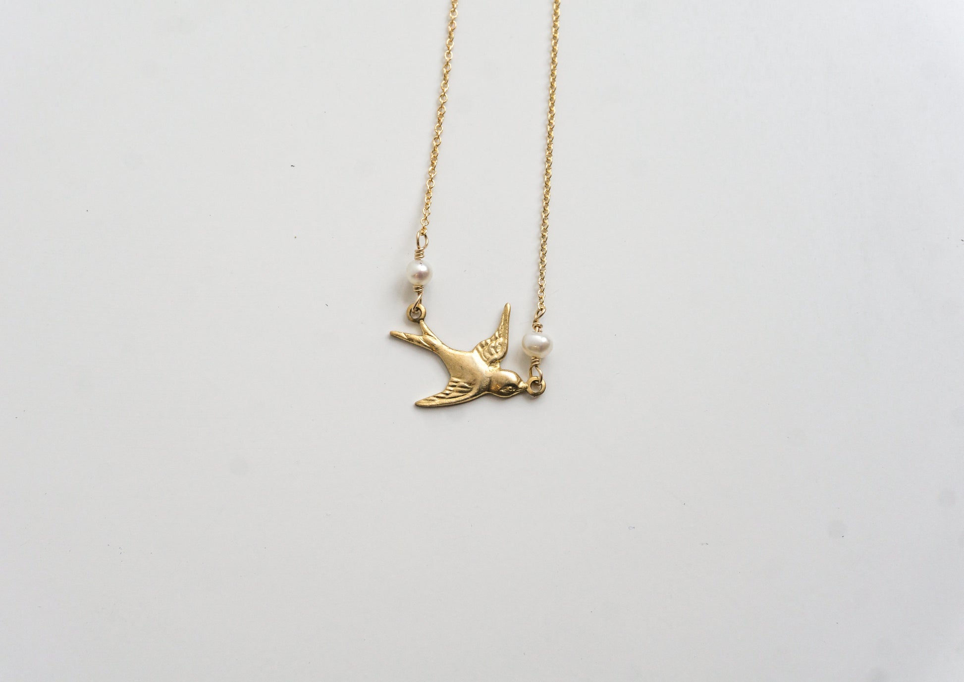 A gold-colored necklace with a charm resembling a bird and two freshwater pearls, displayed against a white background.