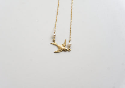 A gold-colored necklace with a charm resembling a bird and two freshwater pearls, displayed against a white background.